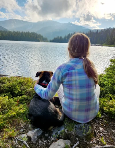 A person sits by a lake, gently holding a dog, with a mountain range and forest in the background under a partly cloudy sky.