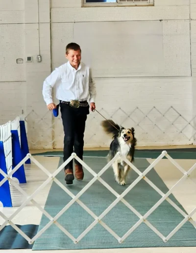 A boy dressed in a white shirt and belt buckle walks alongside a happy dog on a leash during a dog show inside a training facility.