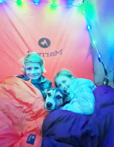 Two children cuddle with a dog in sleeping bags inside a tent, illuminated by colorful string lights.