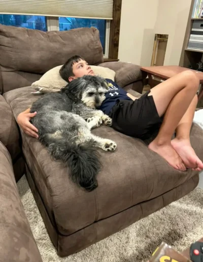 A boy lounges on a couch with a gray and black dog snuggled up against him, both looking relaxed in a cozy living room.