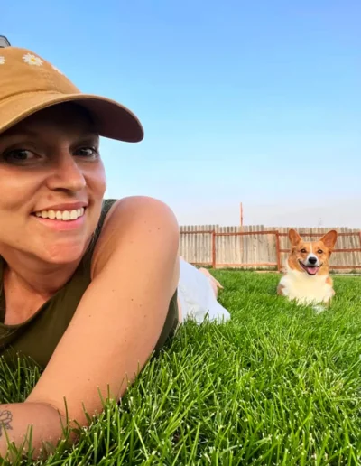 A woman wearing a hat smiles while lying on the grass, with a happy Corgi sitting in the background on the green lawn under a clear sky.