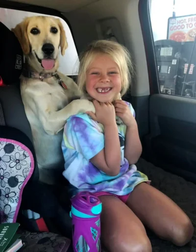 A young girl smiles brightly while sitting in a car next to a happy yellow lab, who playfully places its paws on her shoulders.