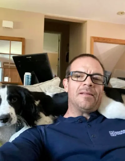 A man wearing glasses and a blue shirt sits on a couch with a black and white dog resting its head and paws on his shoulder.