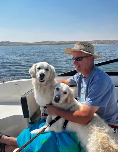 A man in a straw hat and sunglasses sits on a boat, holding two white Golden Retrievers, with calm water and distant hills in the background.