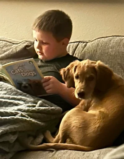 A young boy is sitting on a couch reading a book titled "Cats vs. Dogs" while a light brown puppy sits beside him, looking toward the camera.