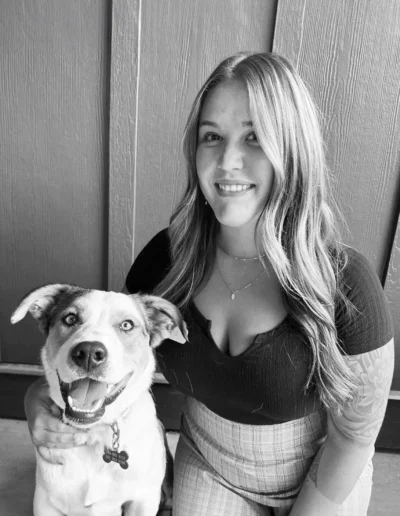 A woman with long wavy hair sits next to a happy dog, both smiling directly at the camera in a black-and-white photo.