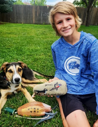 A boy sits on the grass next to a dog, holding a painted rock with the dog's name "Brooks" and a bird illustration on it.