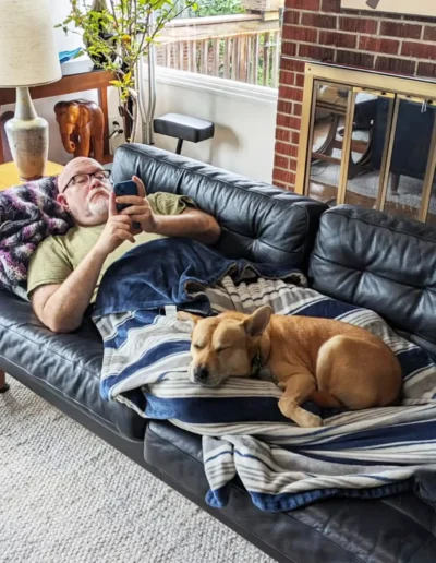 A man relaxes on a black leather couch, looking at his phone, while a tan dog sleeps peacefully beside him, snuggled under a striped blanket. The room has a cozy vibe with warm lighting and a fireplace in the background.